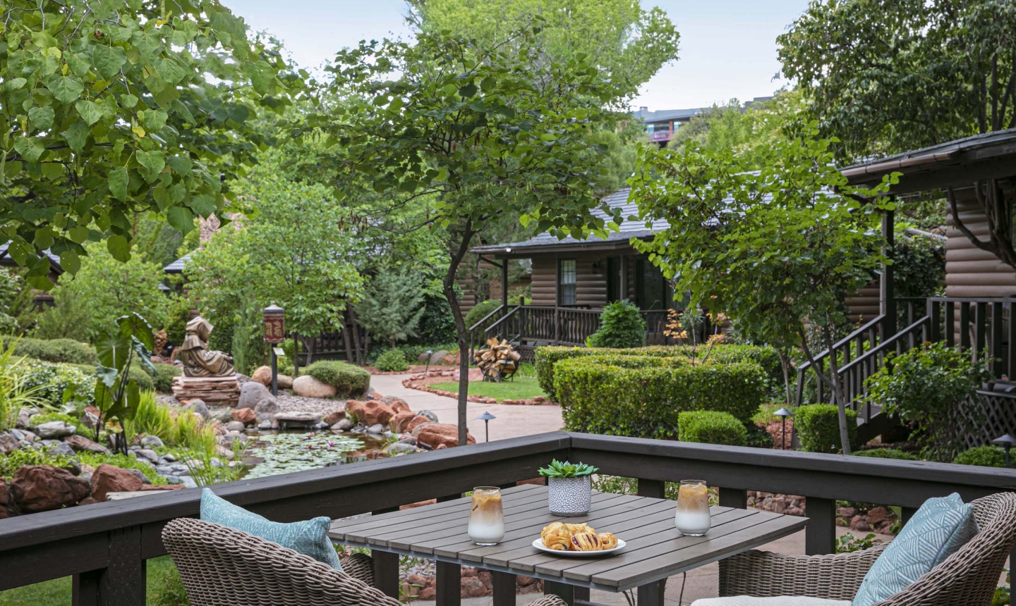View from a cottage deck with a table set for two, overlooking a lush garden with a pond, statues, and log cabins.