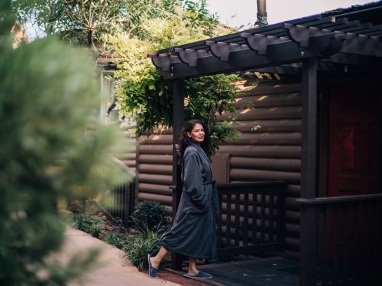 A woman in a dark gray bathrobe smiles as she walks on the wooden deck of a log cabin, surrounded by greenery.