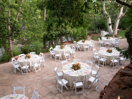 An outdoor wedding reception on a stone patio with round tables and white chairs, set among trees next to a creek.