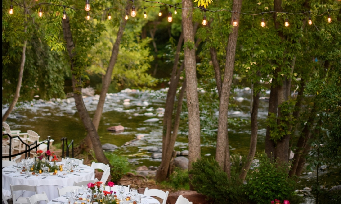 Round tables set for a wedding reception under string lights, on a patio overlooking the flowing water of Oak Creek.