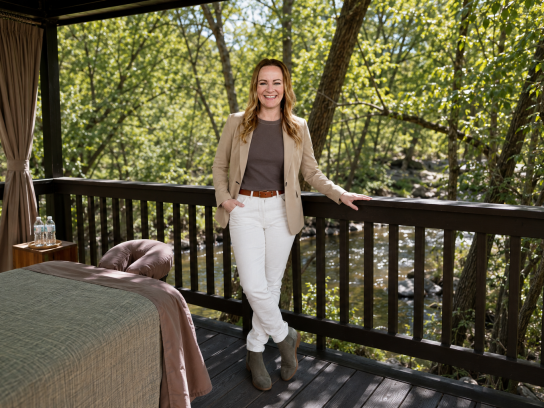 Smiling woman stands on a wooden deck in a forest by a river, wearing a beige blazer, grey top, white pants, brown belt, and grey ankle boots.